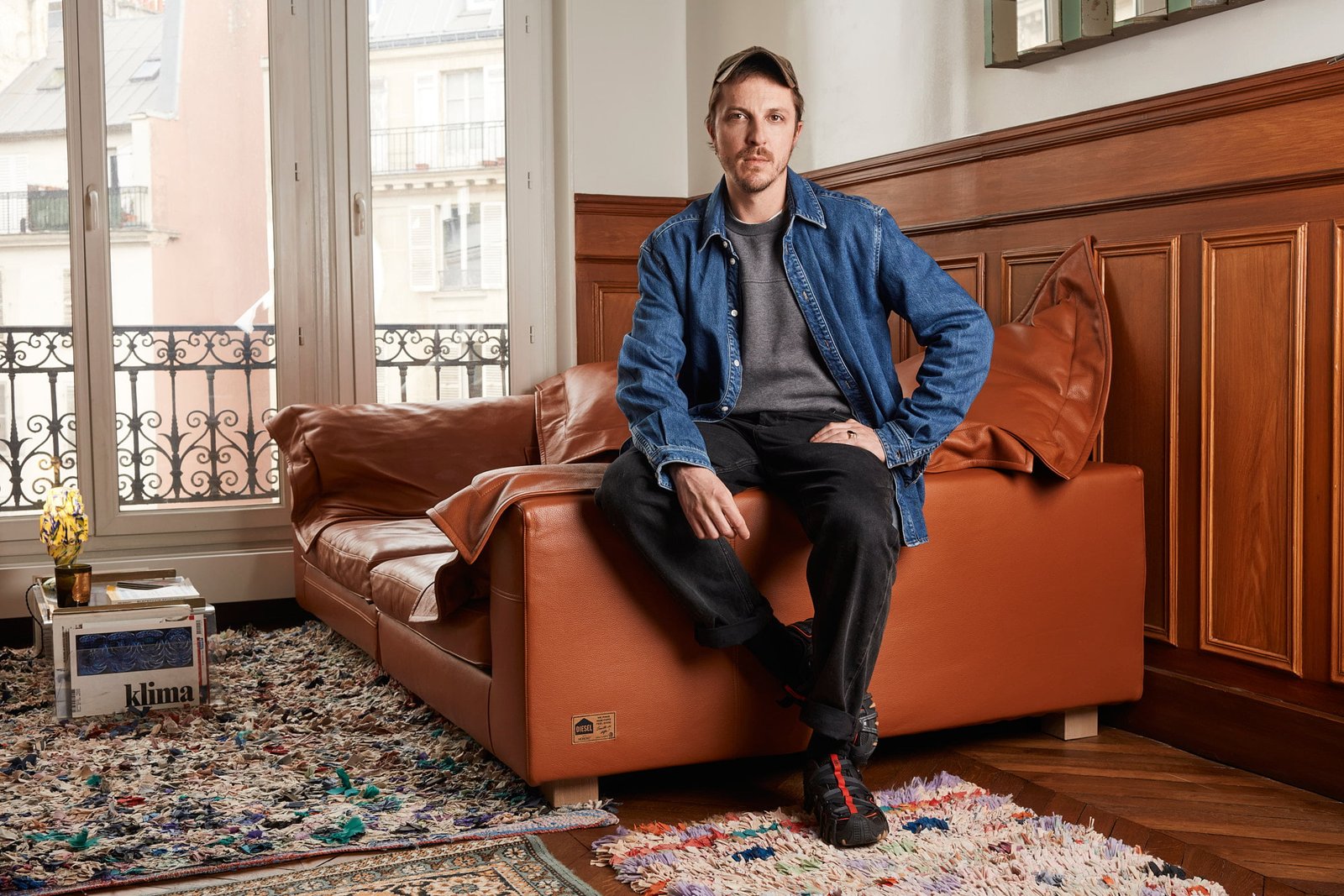 Glenn Martens, directeur artistique de Y/Project, photographiée chez lui, assis sur l'accoudoir de son canapé
